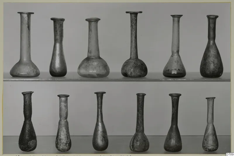 Blown glass vases with long narrow necks, arranged in two rows on shelves against a grey backdrop