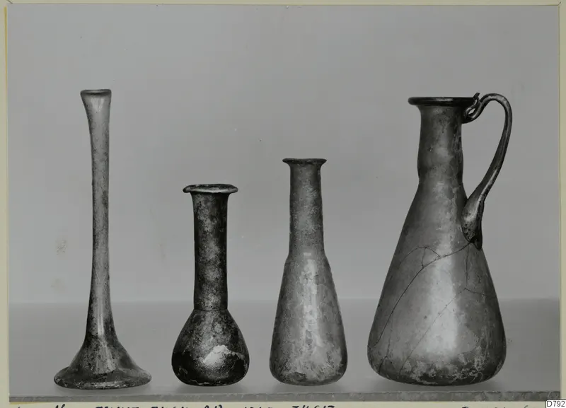 Four blown glass vessels in a row: three slender vases or bottles and a jug with a looped handle.