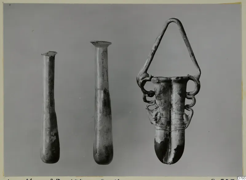 Three glass vessels side by side: two tall slender bottles and one with an arched looped handle.
