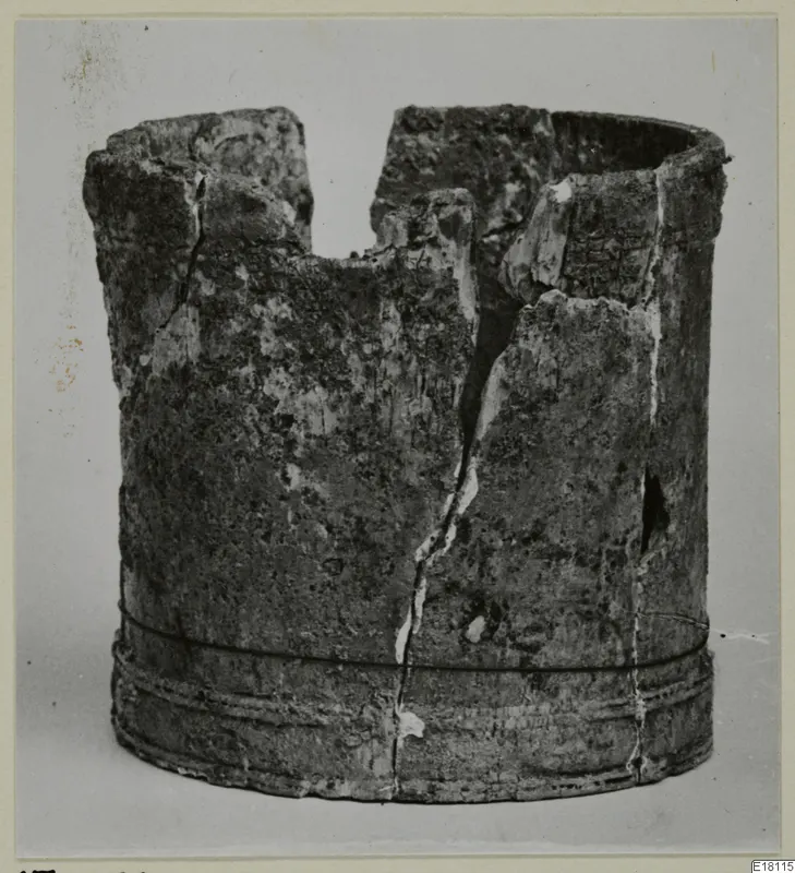 Cylindrical bone object with a broken, uneven rim and grooved bands near the base, photographed against a grey backdrop.