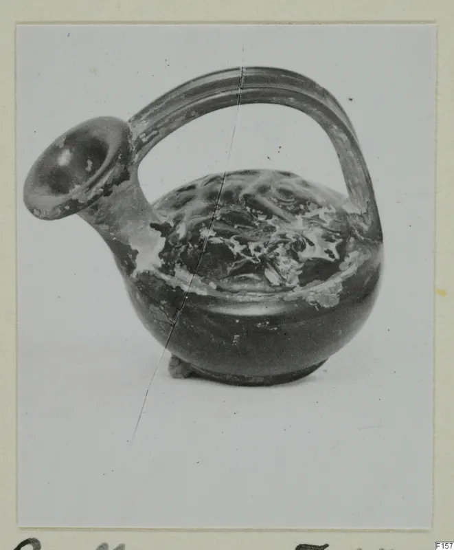 Black glazed vessel with a spout and strap handle, shown on a mounted photograph card