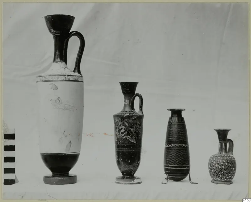 Four vessels in a row on white cloth: a tall white-ground lekythos, a black juglet, a slender banded vessel and a small juglet.