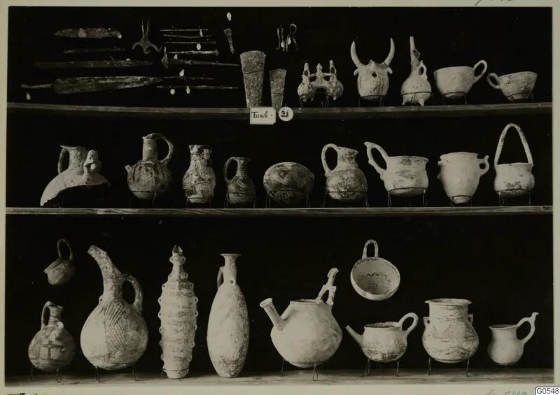 Finds from Lapithos Markides, T.21, on three shelves: metal blades, pins and pottery vessels against a black background.