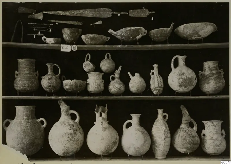 Pottery jugs, bowls and jars with metal blades and pins on three shelves, from T.18 at Lapithos Markides
