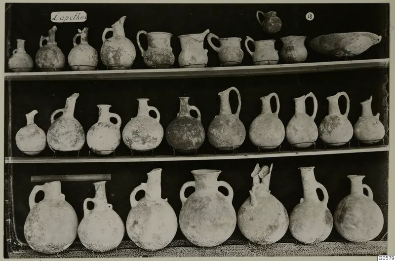 Pottery juglets, jugs and a shallow bowl arranged in three rows on shelves, from Lapithos Markides Tomb 18 A.