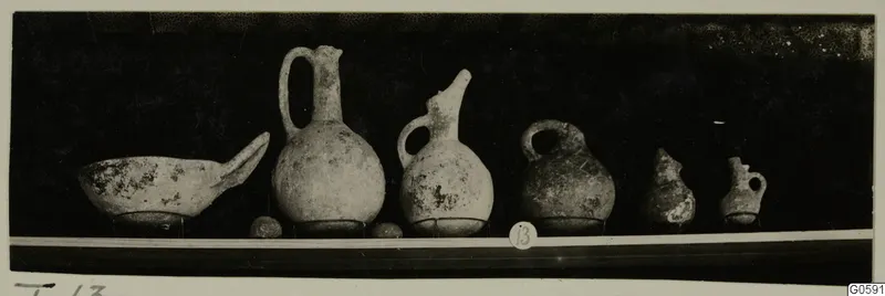 Row of six pottery vessels from Lapithos Markides, T.13, standing on a shelf against a dark background