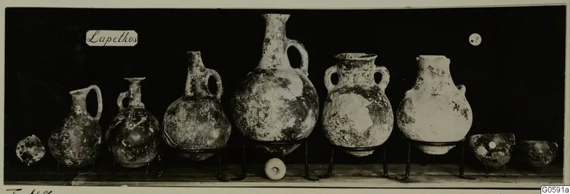 Row of pottery jugs, jars and bowls from Lapithos, Markides tomb 48, set on metal stands against a dark background.