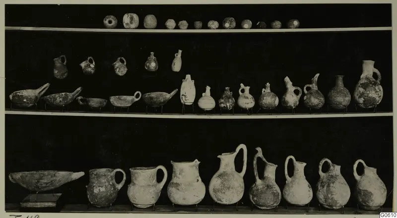 Three shelves of pottery vessels, bowls and jugs from Lapithos Markides, T.49, photographed against a black background.