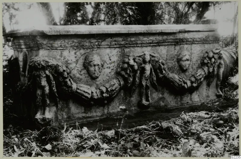 Long side of a stone sarcophagus with carved garland swags and human heads in relief, set among leaves outdoors.