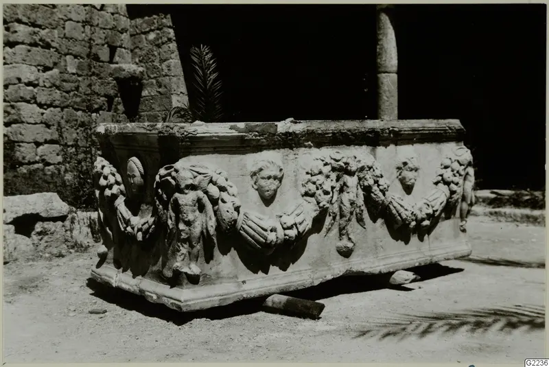 Stone sarcophagus with carved garlands, medallion heads and small figures, standing outdoors by a rubble stone wall.