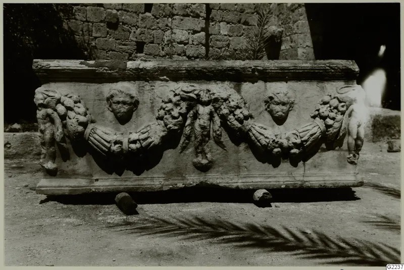 Front of a carved stone sarcophagus with garlands, two carved faces and small figures, standing on dirt before a stone wall