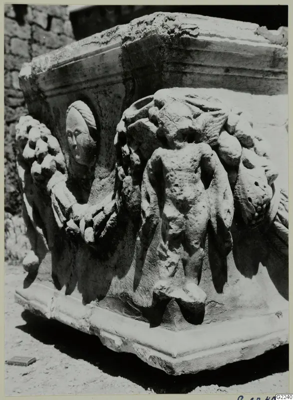 Carved stone block with a head in a round medallion, a garland, and a standing figure in relief.