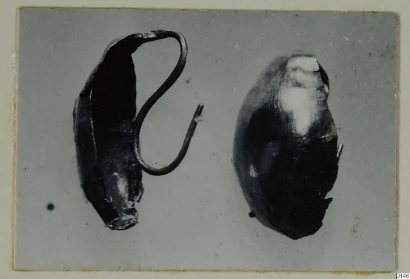 Two dark, corroded metal jewellery pieces side by side: a flattened tube with a bent wire loop, and an oval bead-like object.