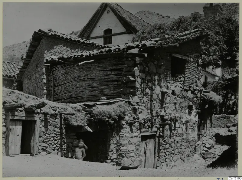 Stone houses with tiled roofs in the village of Lazania, with a stacked reed fence and a person in a doorway