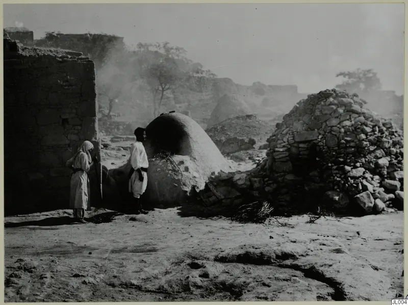 Two people in white robes beside a rounded clay oven and a dome of rough stones, with smoke drifting behind them