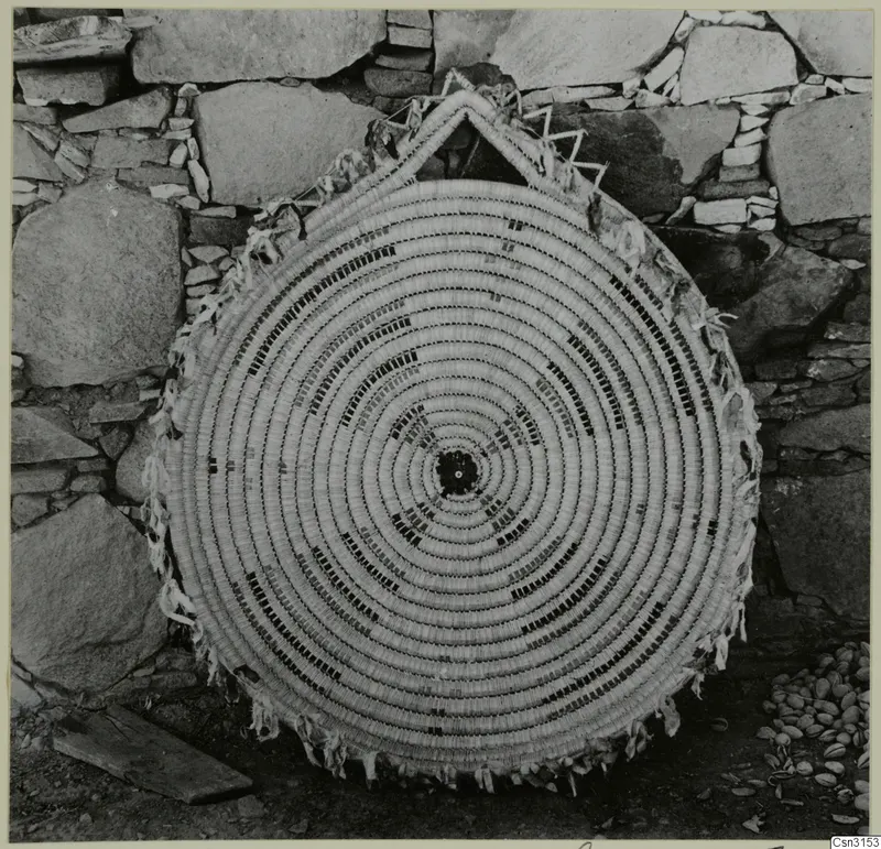 Large circular coiled woven mat with dark patterned bands, leaning against a stone wall