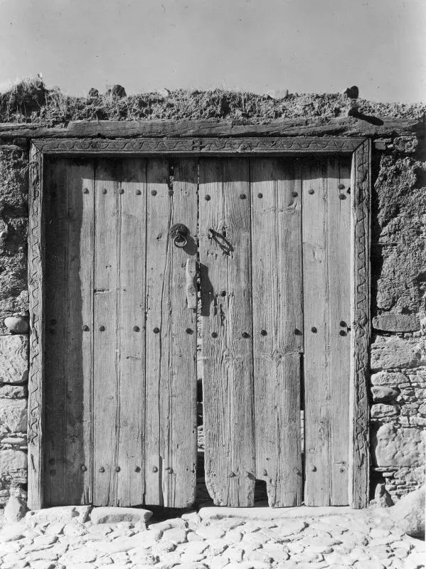 Wooden plank door with studded frame and iron ring knocker in a stone wall, under a thatched roof edge.