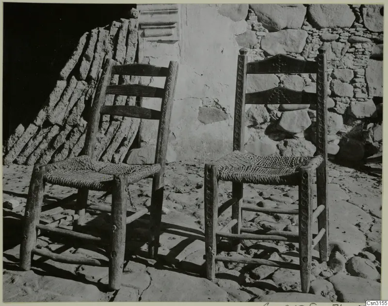 Two wooden chairs with woven seats standing against a stone and plaster wall on a paved ground.