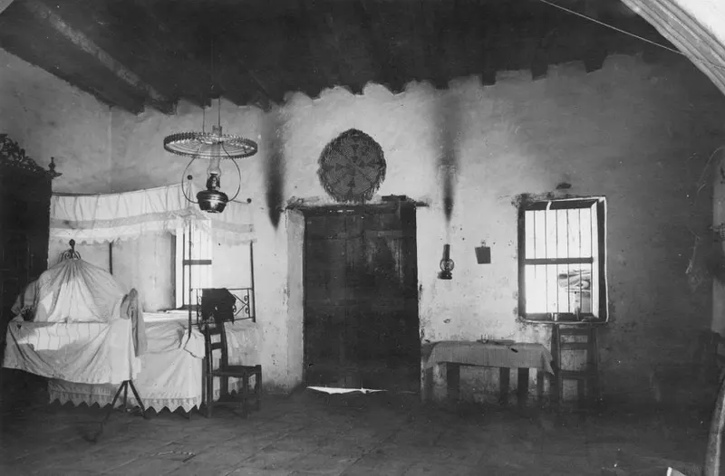 Interior of a whitewashed room with a canopied bed, a hanging oil lamp, a small table and chairs