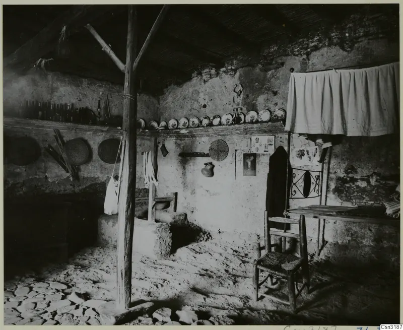 Interior of a plastered room with a wooden roof post, a shelf of plates and bottles, and a woven wooden chair.
