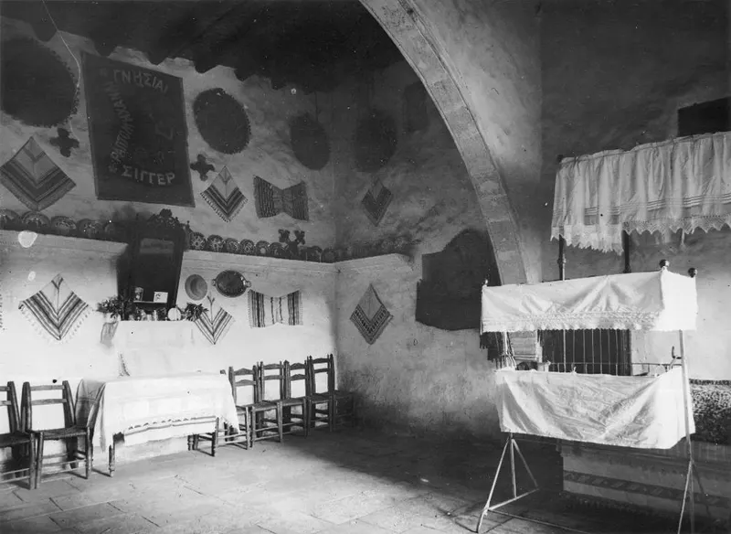 Interior of a vaulted room with textile hangings on the walls, a row of wooden chairs, a covered table and plates on a ledge.