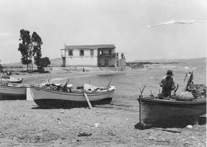 Boats drawn up on a pebbly shore at Boghazi, with a two-storey building and a standing man in a hat