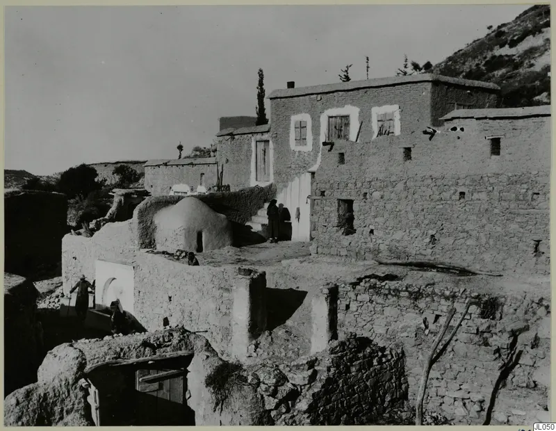 Village houses of mud-brick and stone at Mandres, with people on steps and a low domed structure in the middle