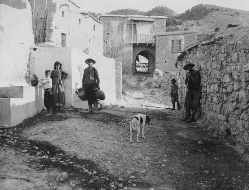 Village street with whitewashed and stone walls, a dog, and villagers with jars and a stick