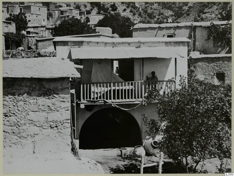 Two-storey building with an arched ground floor and a wooden balcony at Mandres, with a man standing on the balcony.