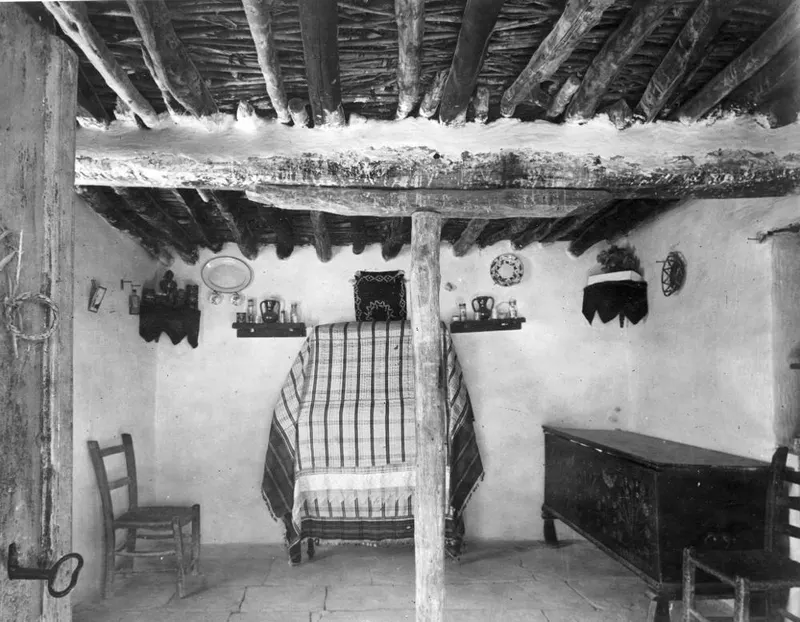 Interior of a plastered room with a timber-beamed ceiling, a striped-covered bed, wall shelves of jugs and a painted chest