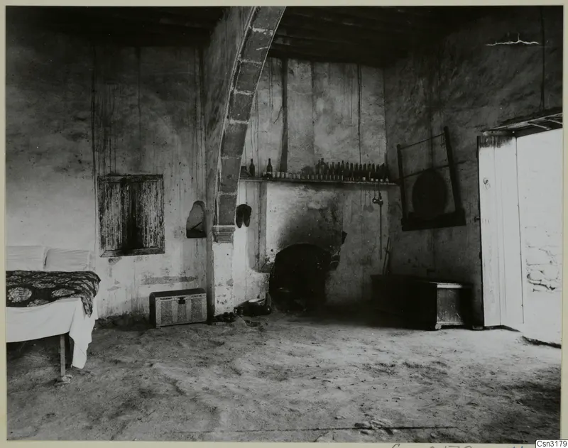 Interior of a plastered room with a shelf of bottles above a hearth, a bed at left and a wooden chest on the floor