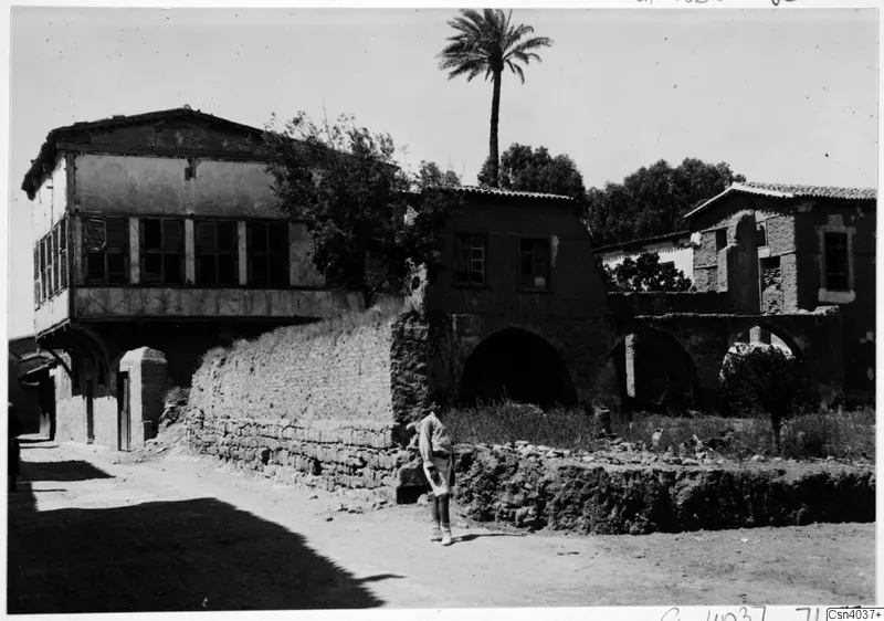 Street in Nicosia with a two-storey house with shuttered upper windows, arched stone walls and a tall palm, with a man standing in the road.