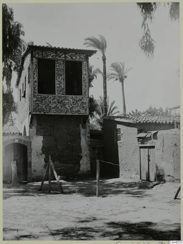 Two-storey building with a timber upper floor of patterned panels, palm trees and a mud-brick annexe beside it, in Nicosia