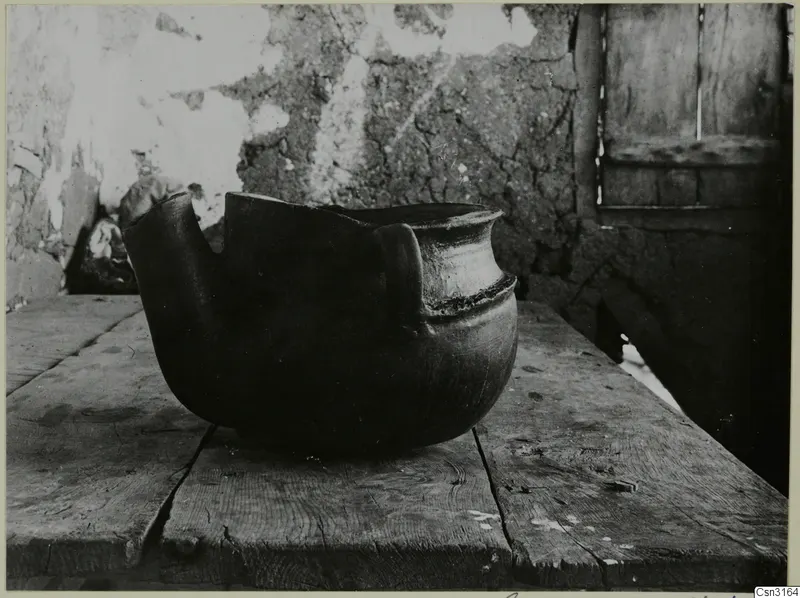 Large round-bodied clay vessel with a handle and broken rim, standing on a wooden table in Klirou