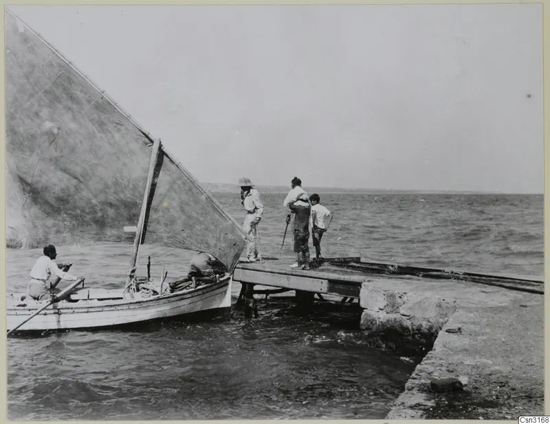 Small sailing boat with a large sail alongside a stone jetty, with several people standing and sitting nearby by the sea.
