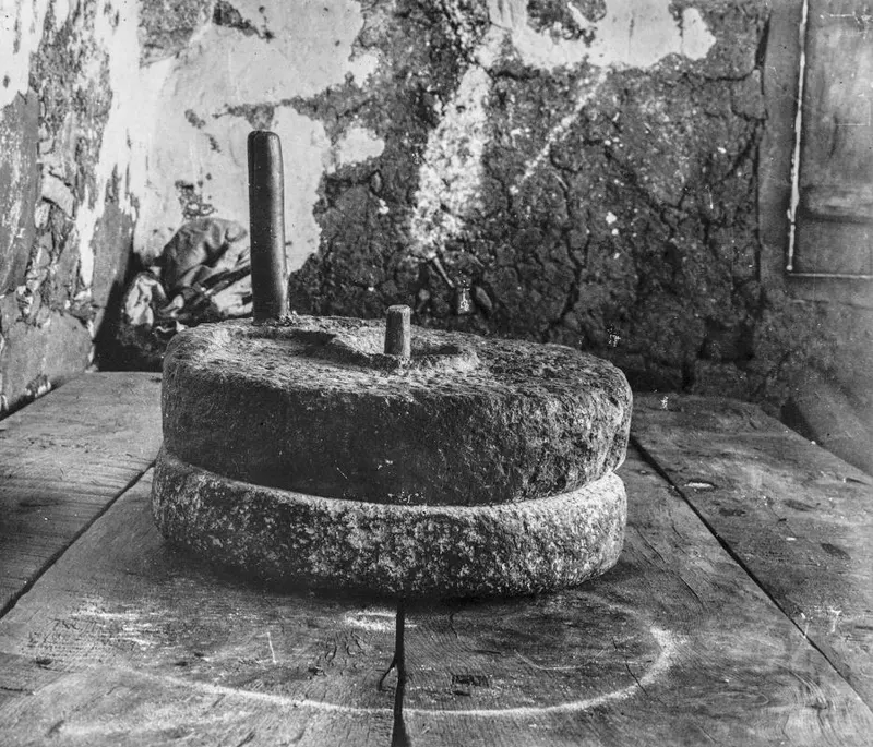 Two-part stone hand mill for grain, with a wooden handle peg, standing on wooden planks