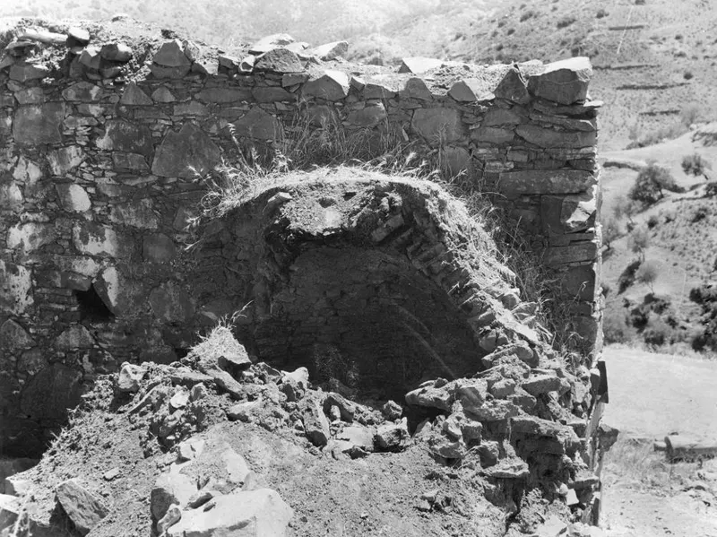 Stone oven with an arched opening set into a rubble-stone wall, with fallen stones and soil in front.