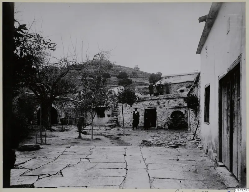 Stone-paved courtyard with a rubble-stone wall, a possible oven recess, and people standing on the wall top