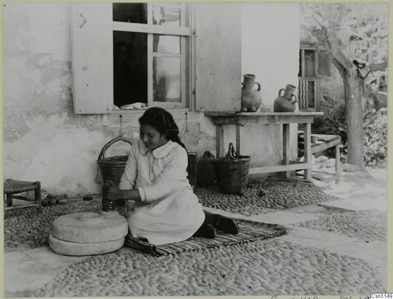 Woman grinding grain with a hand mill on a cobbled yard, with baskets and jugs beside a house wall