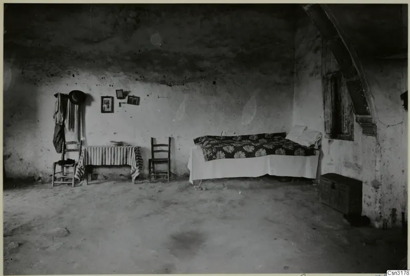 Interior of a whitewashed room with a bed, two wooden chairs and a table covered with striped cloth