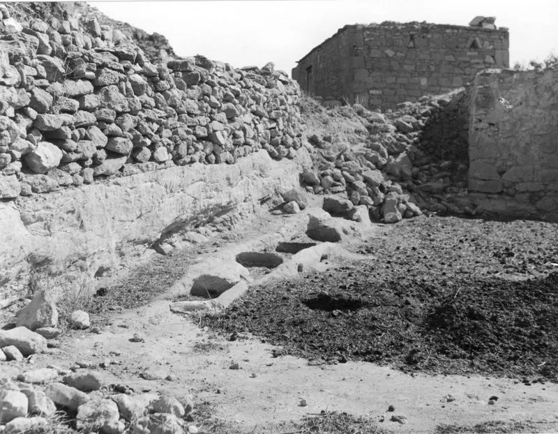Stone feeding troughs cut into a pale ledge beside a rough stone wall at Galinoporni, with a ruined stone building behind.