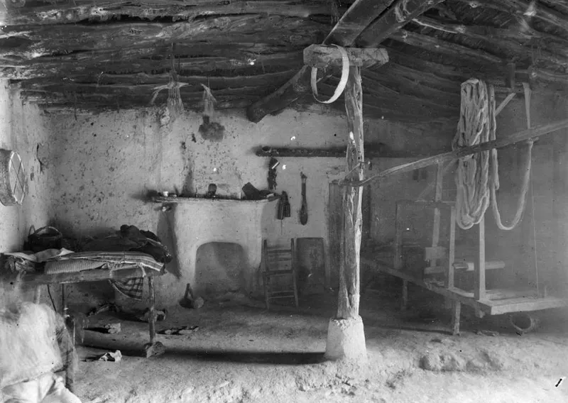 Interior of a plastered room with a branch roof, a wooden loom on the right and a wooden bed on the left.