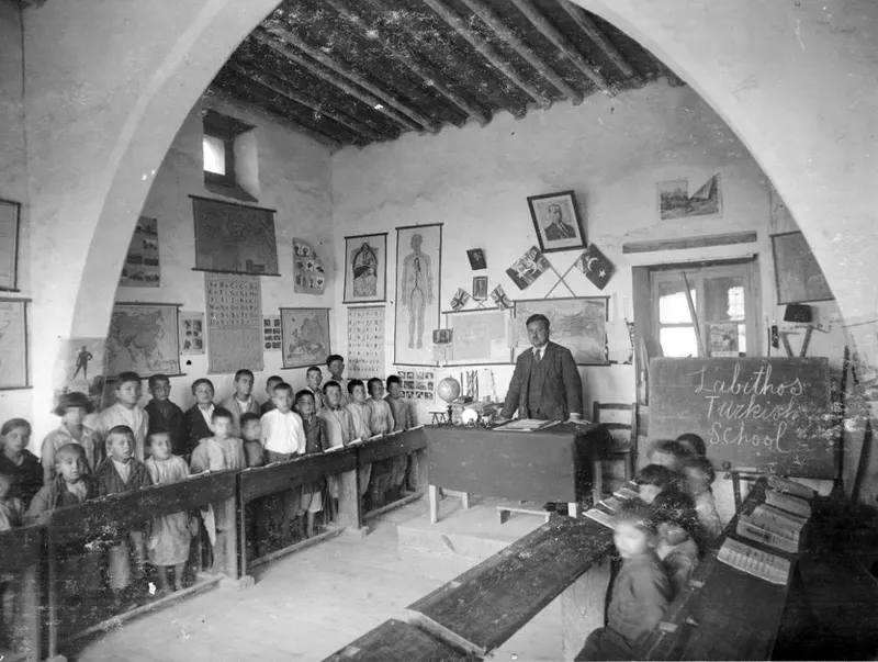 Teacher standing at a desk in an arched whitewashed classroom, with rows of children on benches, Lapithos