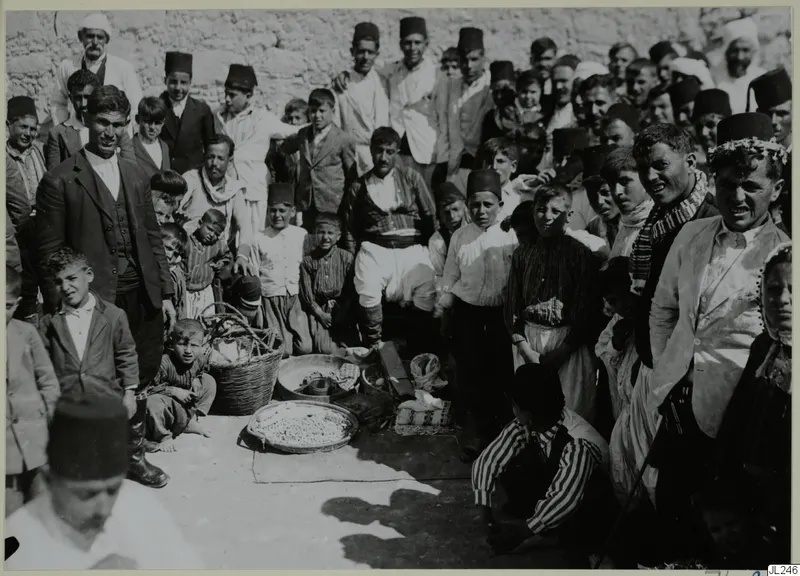 Crowd of men, boys and a woman in fezzes and jackets gathered around baskets and a food tray at Galinoporni.