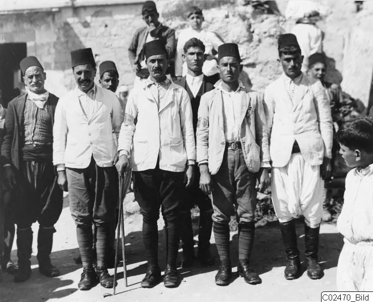 Group photograph of men in light jackets and brimless caps at Ayia Irini, one holding a walking stick