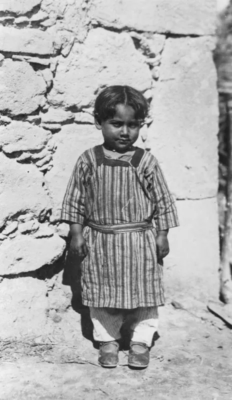 Young child in a striped smock and sash standing in front of a rough stone wall