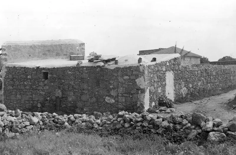 Rough stone structure with a whitewashed flat top beside a dirt track, with a rubble stone wall in the foreground