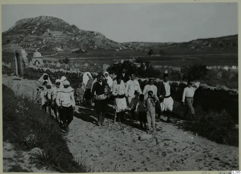 Group of people, women and men with children, walking along a sandy track past a stone wall, with a rocky hill behind.