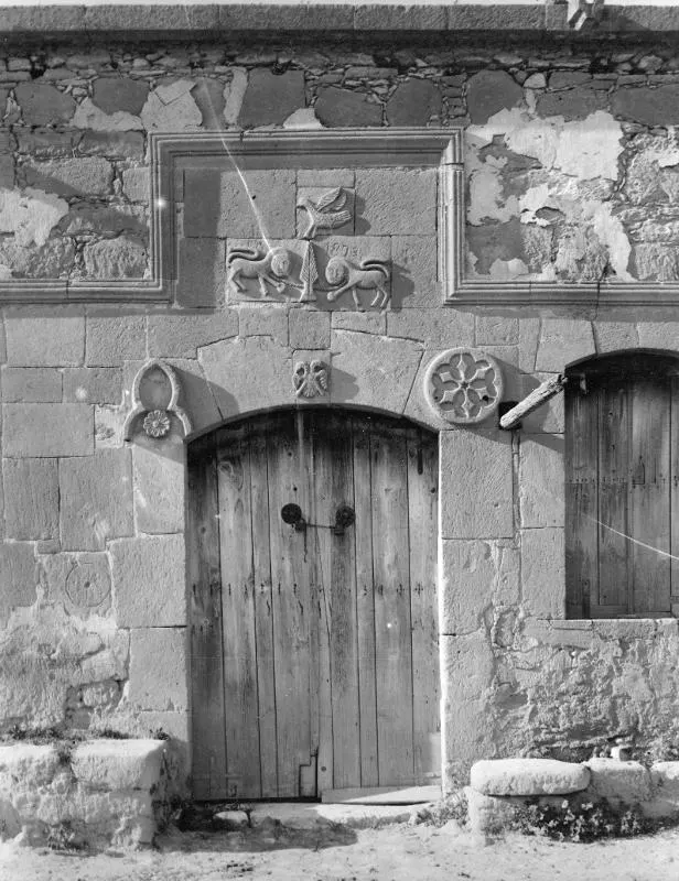 Stone building front with a wooden plank door under an arch, carved reliefs above it and a rosette medallion beside the door.