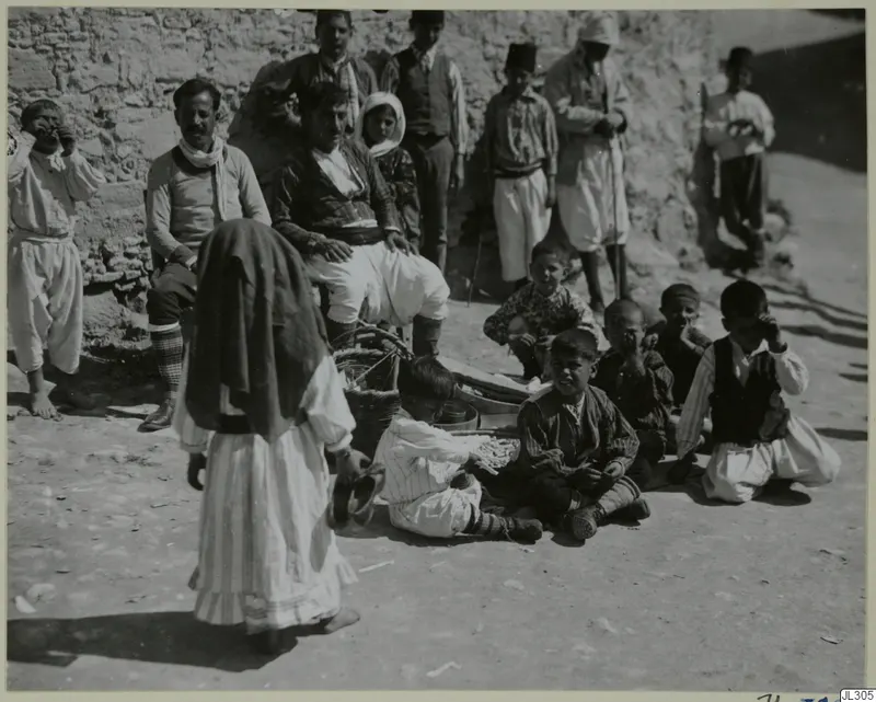 Group of children and men in front of a rough stone wall, with a woman in a dark head shawl walking in the foreground.